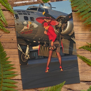 Puede incluir: Una chica pin-up con un vestido rojo y medias negras se encuentra frente a un avión vintage. El avión está pintado de plata y tiene un diseño rojo, blanco y azul en el lateral. La chica sostiene una sombrilla roja y mira a la cámara.