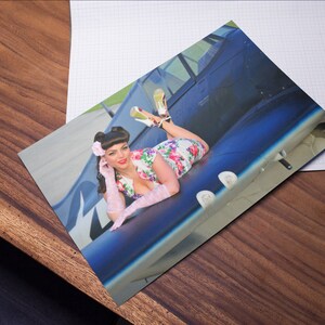 May include: A woman in a floral dress and white gloves poses on the wing of a blue airplane. She is wearing a pink flower in her hair and has a playful expression on her face.
