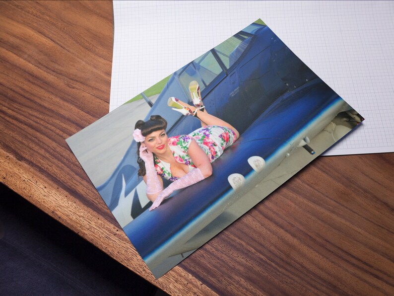 May include: A woman in a floral dress and white gloves poses on the wing of a blue airplane. She is wearing a pink flower in her hair and has a playful expression on her face.