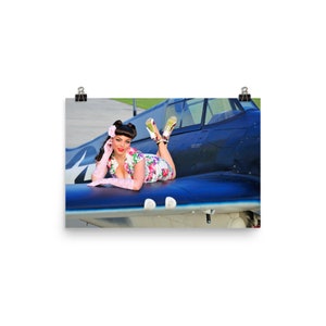 May include: A woman in a floral dress and pink gloves lies on the wing of a blue and white airplane. She is wearing pink heels and has a pink flower in her hair.