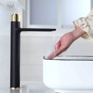 Op de afbeelding: Een moderne zwarte kraan met gouden accenten. De kraan is hoog en slank, met een lange uitloop. Water stroomt uit de uitloop in een witte wastafel. De basis en de bovenkant van de kraan zijn goudkleurig.
