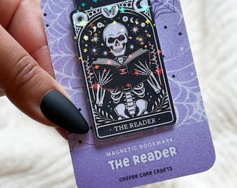 May include: A magnetic bookmark featuring a black and white illustration of a skeleton reading a book, with the text "THE READER." The bookmark is attached to a purple card with spiderweb and moon designs. The text "MAGNETIC BOOKMARK The Reader coffee cake crafts" is also on the card.