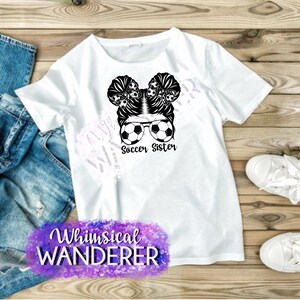 May include: White t-shirt with a black and white graphic of a girl with soccer balls in her hair and sunglasses. The text "Soccer Sister" is below the graphic. The text "Whimsical Wanderer" is in purple and pink at the bottom of the shirt.