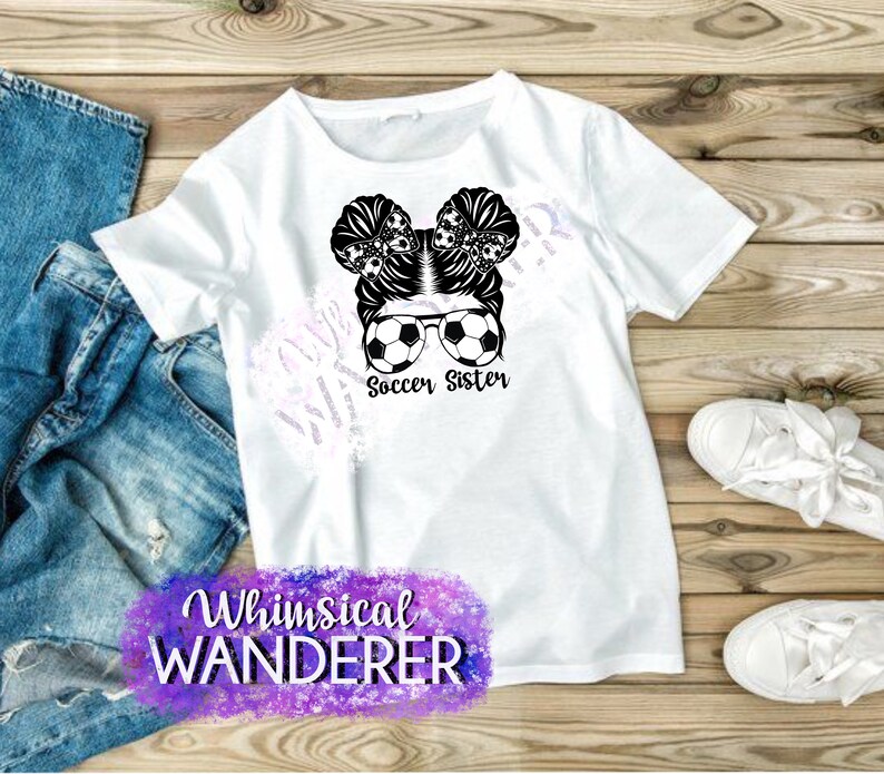 May include: White t-shirt with a black and white graphic of a girl with soccer balls in her hair and sunglasses. The text "Soccer Sister" is below the graphic. The text "Whimsical Wanderer" is in purple and pink at the bottom of the shirt.