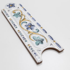 Puede incluir: Azulejo de cer&aacute;mica blanco con un dise&ntilde;o floral decorativo en tonos azules, verdes y dorados. El azulejo tiene un borde elevado y un borde festoneado.