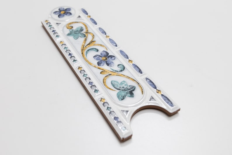 Puede incluir: Azulejo de cer&aacute;mica blanco con un dise&ntilde;o floral decorativo en tonos azules, verdes y dorados. El azulejo tiene un borde elevado y un borde festoneado.