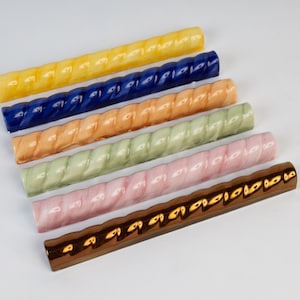 May include: A collection of six ceramic, twisted-design bars in various colors. The colors include yellow, blue, orange, green, pink, and brown. The bars are arranged parallel to each other on a white background. Each bar has a glossy finish.