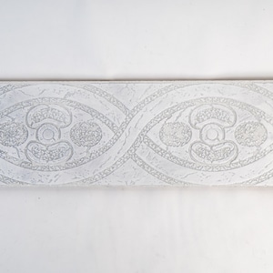 May include: A rectangular, off-white decorative tile with a repeating pattern. The design features interwoven oval shapes with floral and circular motifs, all in a raised, textured relief. The tile is set against a plain white background.