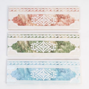May include: Three ceramic tile border pieces with a white background and a decorative pattern. The top tile is red with a white floral design, the middle tile is green with a white floral design, and the bottom tile is blue with a white floral design.