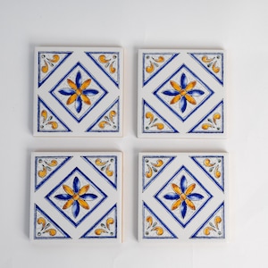 May include: Four white ceramic tiles with a blue and yellow floral design. Each tile features a diamond shape with a stylized flower in the center.