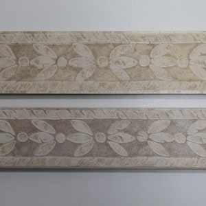 May include: Two beige ceramic tile border pieces with a white floral design. The design features a repeating pattern of stylized leaves and dots.