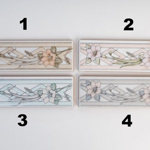 May include: Four ceramic tile samples with a floral design. The tiles are arranged in two rows of two. The top row has a light beige tile with a pink flower and a light pink tile with a pink flower. The bottom row has a light blue tile with a white flower and a light gray tile with a white flower.