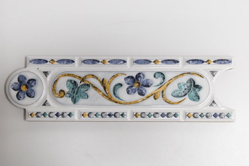 Puede incluir: Azulejo de cer&aacute;mica blanco con un dise&ntilde;o floral azul y verde. El azulejo tiene un borde dorado y un borde de diamantes azules y blancos.