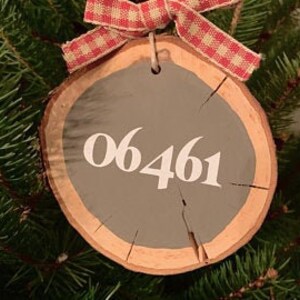 Op de afbeelding: Een ronde houten kerstversiering met een grijs geschilderd midden en het nummer "06461" in wit. De versiering heeft een natuurlijke houtnerfrand en is versierd met een rood-wit geruit lint om op te hangen.