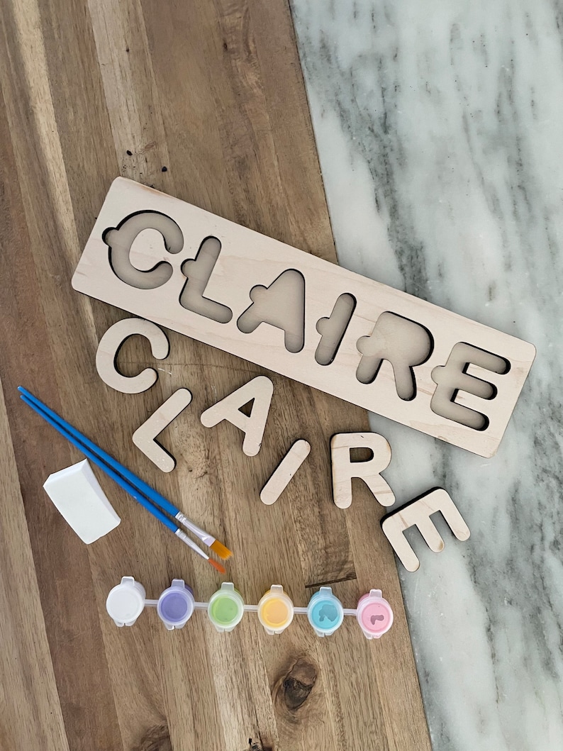 May include: Wooden name puzzle with the name "CLAIRE" cut out in large letters. The puzzle pieces are scattered around the base. The set includes six small paint pots in various colors, two paintbrushes, and a sponge.