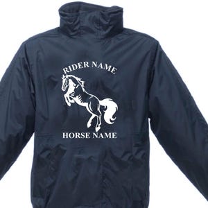May include: Dark navy equestrian jacket featuring a white horse illustration and the text "RIDER NAME" and "HORSE NAME". The jacket has a high collar and a zip. A practical garment for horse riding.
