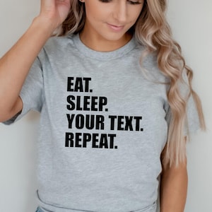 以下が含まれることがあります： 「EAT. SLEEP. YOUR TEXT. REPEAT.」という太字の黒い文字がプリントされた、明るい灰色のTシャツ。クルーネックのスタイルで、柔らかい素材で作られているようです。