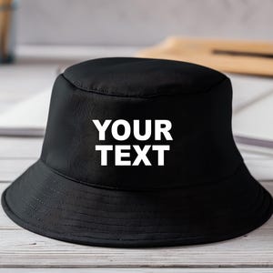 Op de afbeelding: Een effen zwarte bucket hat met een platte bovenkant en een naar beneden lopende rand. Op de voorzijde van de hoed staat de tekst YOUR TEXT in dikke, witte, schreefloze hoofdletters, duidelijk gecentreerd op de donkere stoffen ondergrond.