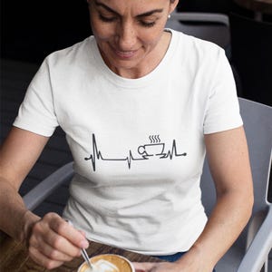 May include: A white t-shirt featuring a black graphic of a coffee cup with steam rising from it, enclosed within a heartbeat line. The text "I love coffee" is not visible in the image. A person is wearing the shirt and holding a cup of coffee.