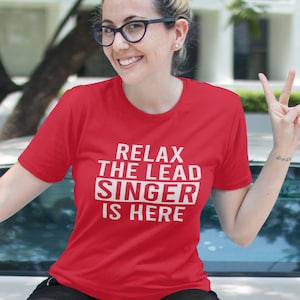 May include: A red t-shirt with the white text "RELAX THE LEAD SINGER IS HERE". The short-sleeved shirt has the text in the centre. The person wearing the shirt is smiling and wearing glasses.