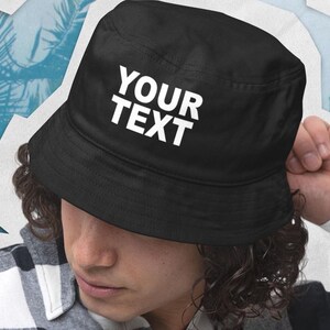 Op de afbeelding: Zwarte vissershoed met de tekst "YOUR TEXT" in witte blokletters. De hoed wordt gedragen door een persoon met krullend haar en een geruit overhemd. De achtergrond heeft een blauw en wit tropisch ontwerp.