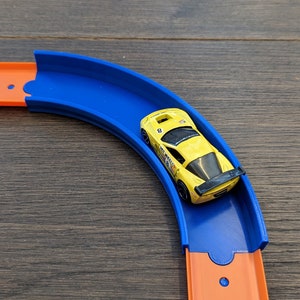 Op de afbeelding: Een gele speelgoedauto met een zwarte spoiler en het nummer 8 aan de zijkant rijdt over een blauw en oranje plastic circuit.