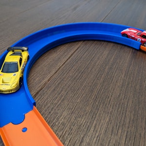 Pode incluir: Um carro de corrida de brinquedo amarelo em uma pista azul e laranja. O carro está na parte azul da pista, que é curva. A parte laranja da pista é reta e leva à parte azul.