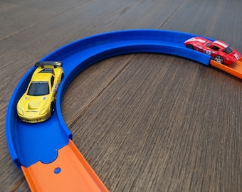 May include: A yellow toy race car on a blue and orange track. The car is on the blue part of the track, which is curved. The orange part of the track is straight and leads to the blue part.