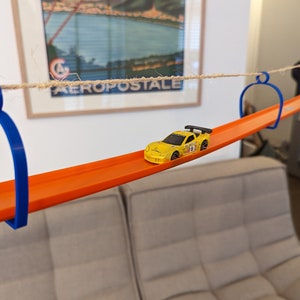 Op de afbeelding: Een gele speelgoedauto racet over een oranje baan die wordt opgehangen door blauwe plastic lussen en een touw. De baan hangt boven een grijze bank.