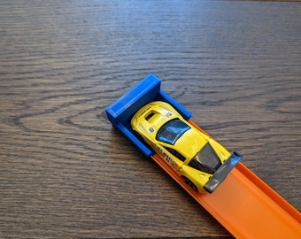 May include: A yellow toy race car with black accents sits in a blue starting gate, positioned on an orange track. The track is set on a wooden surface. The car has the number 8 on it.