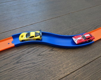 May include: A yellow toy car with a black and white racing stripe and a red toy car with a white stripe are on a blue and orange toy race track.