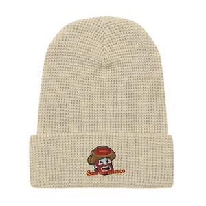 May include: A beige waffle knit beanie with an embroidered cartoon character wearing a red hat and a red beard. The character is holding a red sign that says "San Fran". The text "San Francisco" is embroidered below the character.
