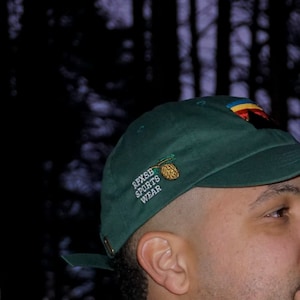 May include: A green baseball cap with a white embroidered logo that says "RPXSB SPORTS WEAR" and a small embroidered pine cone design. The cap has a curved brim and a colorful patch on the back.