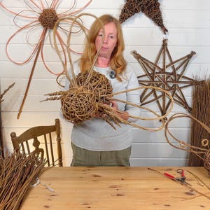 Puede incluir: Una mujer sostiene una cesta tejida a mano con ramitas marrones naturales. La cesta tiene forma esférica y asas largas y curvas. En el fondo se ven otras decoraciones tejidas, como una estrella y una flor.