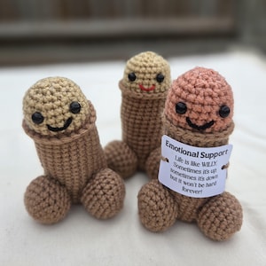 May include: Three phallic-shaped crochet figures with smiling faces and black bead eyes. Two figures are tan and brown, while one is pink and brown. A white tag on the front reads: Emotional Support. Life is like WILLY. Sometimes it's up, sometimes it's down but it won't be hard forever!