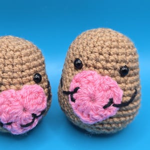 May include: Two handmade crochet mole toys. Each mole has a brown body, black eyes, and a pink nose. The toys are set against a bright blue background. The crochet stitches are clearly visible.