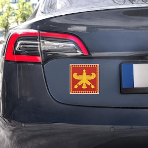May include: A red and gold car sticker featuring a stylized Faravahar symbol, a Zoroastrian emblem. The sticker is square with a red border and a central design of a winged figure with a sun disc and a flowing tail. The sticker is affixed to the rear of a dark gray car.