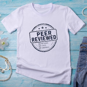 Può includere: Una maglietta bianca con scollo tondo con un design a timbro circolare che recita "PEER REVIEWED". Il timbro include le parole "METHODS", "STATS" e "CONTROLS". La maglietta è abbinata a jeans blu, gioielli e fiori.