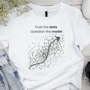 Può includere: T-shirt bianco con la scritta "Trust the data Question the model" e una grafica a freccia grigia. La maglietta è abbinata a jeans blu, scarpe da ginnastica bianche e cuffie nere.
