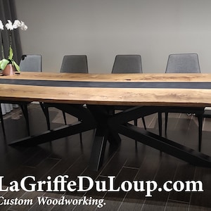 Peut inclure: Une longue table à manger en bois avec un insert en résine époxy noire. La table a un piétement en métal noir avec un design en X unique. La table est dressée avec six chaises et se trouve dans une salle à manger moderne.