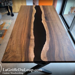 Puede incluir: Una mesa rectangular de madera con un río de resina epoxi oscura en el centro. La mesa es de madera marrón oscuro y tiene una base de metal negro. La mesa mide aproximadamente 183 cm de largo.