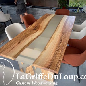 May include: A long wooden dining table with a gray epoxy resin inlay. The table is surrounded by four chairs, two with white upholstery and two with orange upholstery.