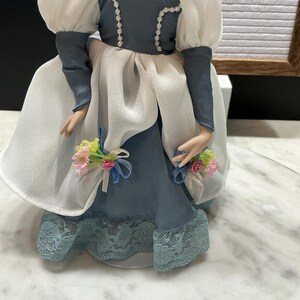 May include: A porcelain doll in a blue and white dress with a lace hem. The doll has a pearl necklace and a blue bodice with white trim. The doll's sleeves are white and puffy. Small bouquets of pink and yellow flowers are attached to the doll's dress.