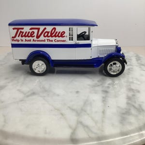 May include: A die-cast model of a vintage True Value delivery truck. The truck is white with a blue roof and blue accents. The True Value logo and slogan are printed on the side. The truck has chrome wheels.
