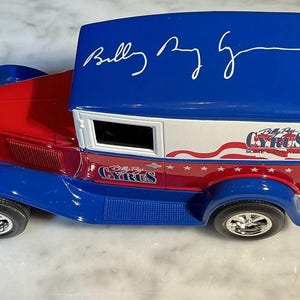 May include: A die-cast model of a vintage delivery truck in red, white, and blue. The truck features the text "Billy Ray Cyrus" and a signature on the roof. The side of the truck has an American flag design.