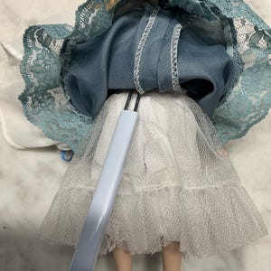 May include: A vintage doll with blonde hair, wearing a blue dress with a lace trim and a white tulle skirt. The doll is supported by a light blue stand.