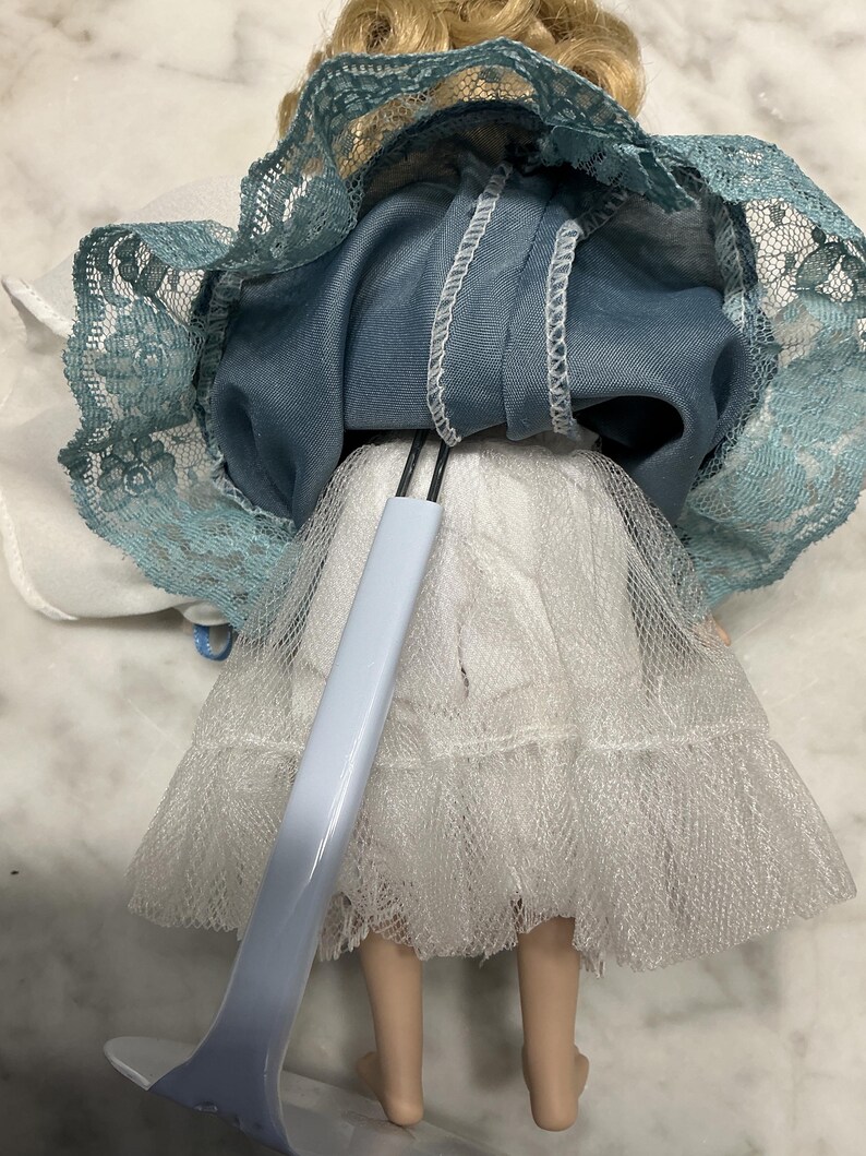 May include: A vintage doll with blonde hair, wearing a blue dress with a lace trim and a white tulle skirt. The doll is supported by a light blue stand.