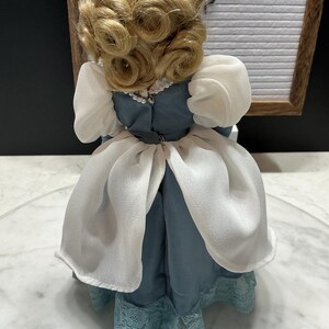 May include: A vintage doll with blonde, curled hair, wearing a blue dress with white accents and a lace hem. The doll's dress has a high neckline and puffed sleeves. The doll is a collectible item.