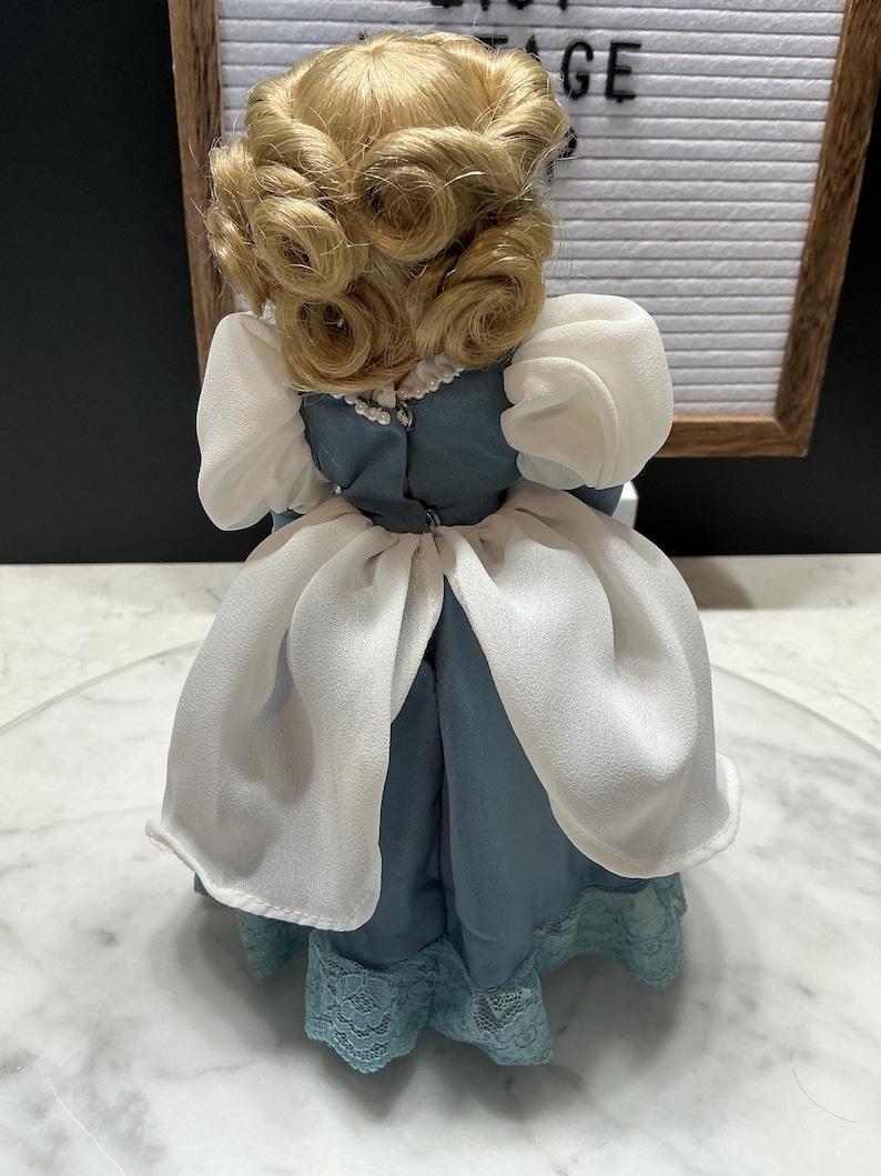 May include: A vintage doll with blonde, curled hair, wearing a blue dress with white accents and a lace hem. The doll's dress has a high neckline and puffed sleeves. The doll is a collectible item.