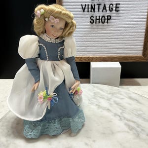 May include: A vintage porcelain doll with blonde hair and a blue dress with white accents. The doll has a floral headpiece and is holding a bouquet of flowers. The background includes a sign that reads "BMKTREASURES ETSY VINTAGE SHOP."
